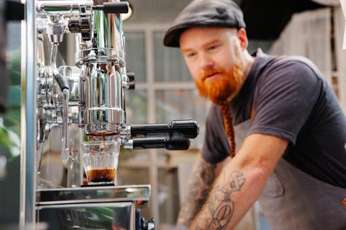 Hipster barista against coffee maker brewing espresso in kitchen