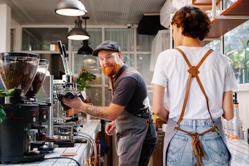 Cheerful bearded male cafe employee talking to anonymous female colleague against professional coffee maker and grinders