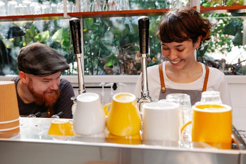 Content bearded male employee talking to smiling young female coworker against coffee maker with bright mugs in cafeteria kitchen