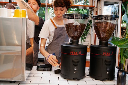 Crop female worker with brush near hipster male partner against professional electric coffee maker and grinders in cafeteria