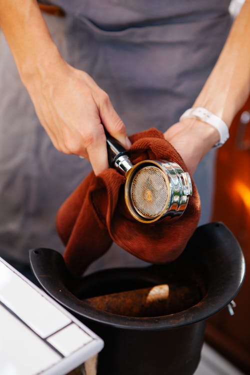 Crop anonymous female cafeteria worker cleaning stainless steel portafilter above coffee grinder at work