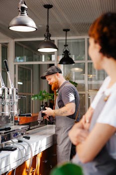 Side view of smiling unshaven male cafe worker talking to crop unrecognizable female partner against professional coffee machine