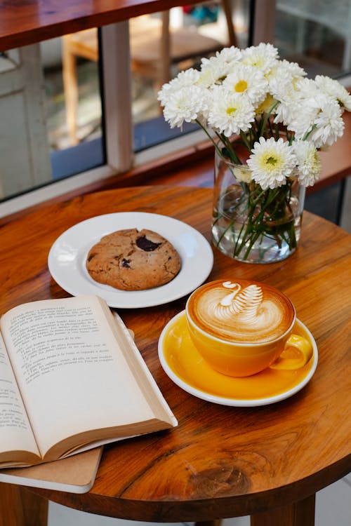 Free From above of tasty coffee with ornament on froth near biscuit and open book on cafeteria table Stock Photo