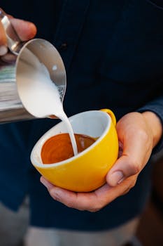 Crop anonymous male barista pouring hot frothy milk into yellow cup with aromatic latte coffee in modern cafeteria