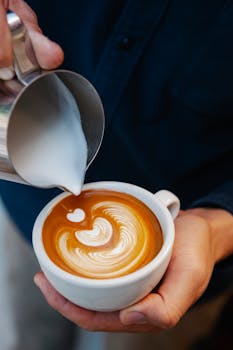 High angle crop unrecognizable male barista adding fresh frothy milk from pitcher into cup of latte and making latte art