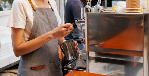 Crop anonymous female in apron making hot coffee with special coffee machine in kitchen of cafe