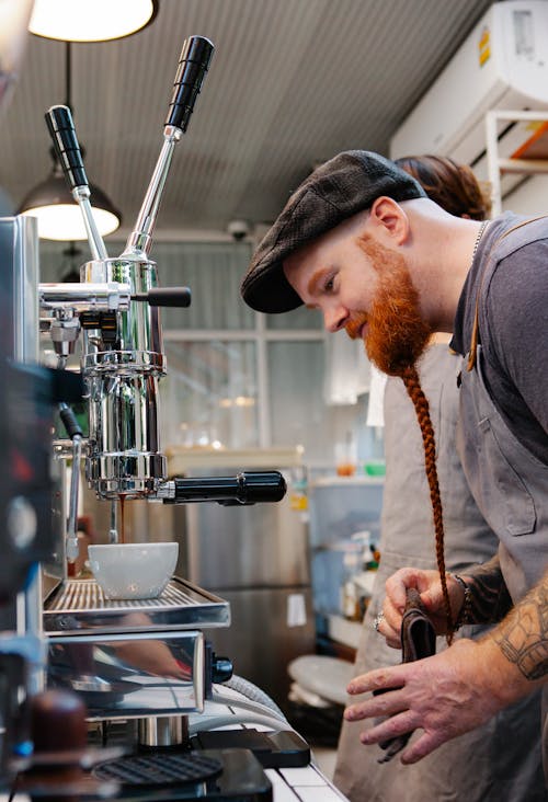 Free Side view of happy young bartender with red beard with pigtail making coffee with coffee machine Stock Photo