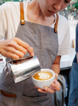 Crop female in apron preparing cup of coffee with latte art and warm milk in cafe