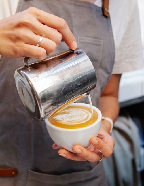 Free Crop anonymous female in apron pouring cream in mug with coffee with latte art Stock Photo