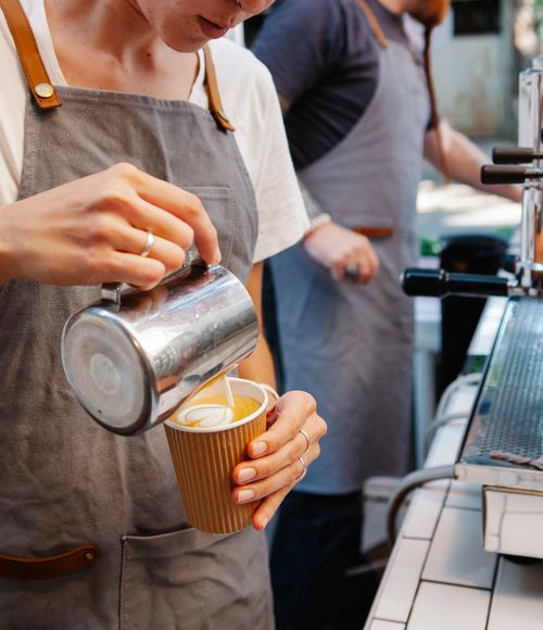 Free Crop anonymous female in apron pouring cream from pitcher into cup with fragrant coffee Stock Photo