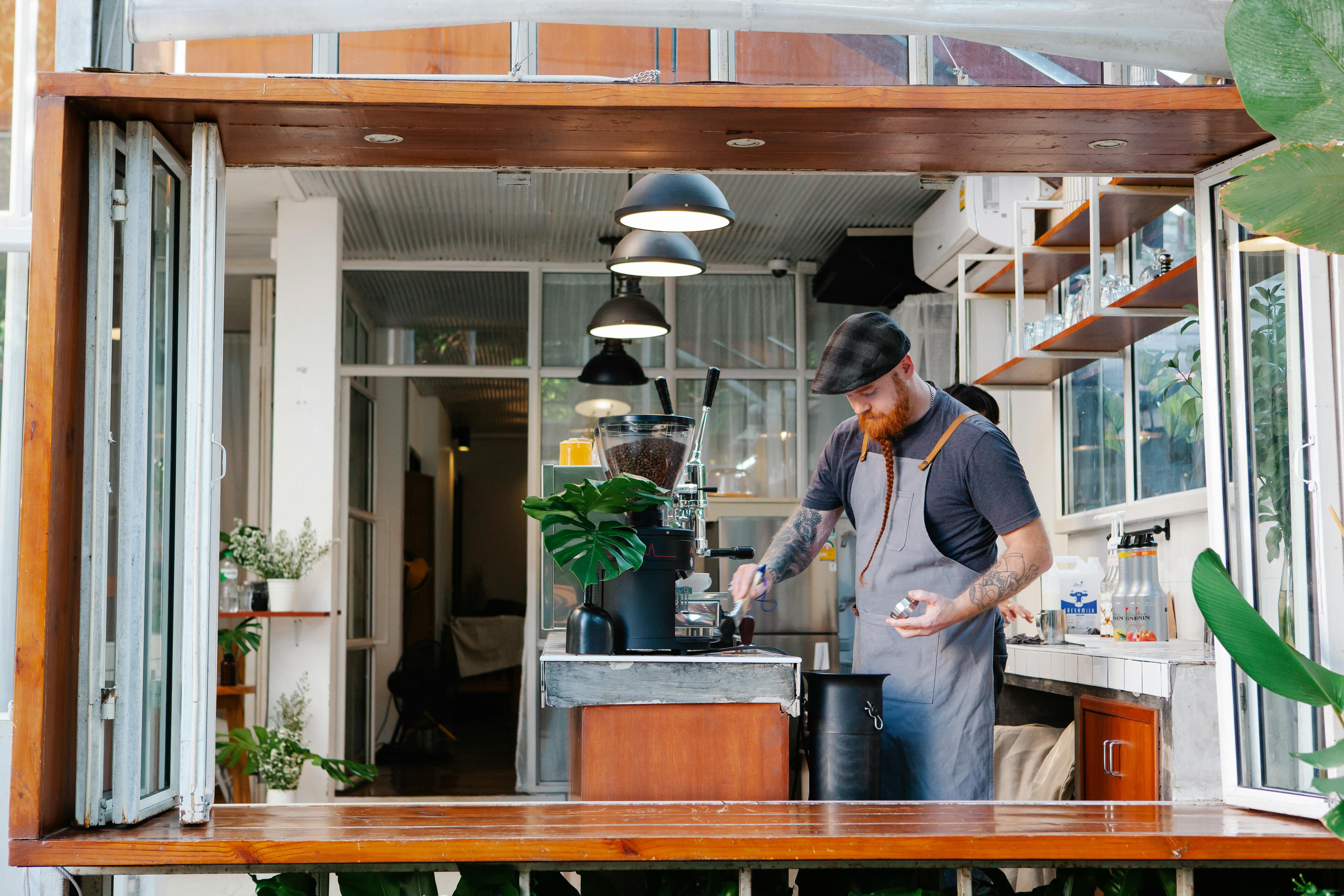 Pensive young tattooed male with pigtail in red beard using coffee machine in kitchen with lamps by Tim Douglas