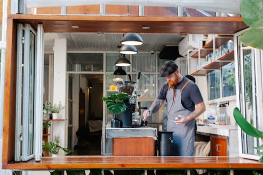 Pensive young tattooed male with pigtail in red beard using coffee machine in kitchen with lamps