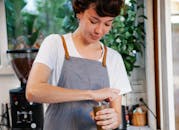 Crop happy female in apron smiling and using grinder for preparing aromatic coffee in kitchen