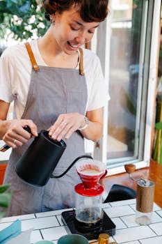 Crop cheerful female barista in casual clothes and apron brewing fresh coffee with filter dripper in light kitchen in cafe in daytime