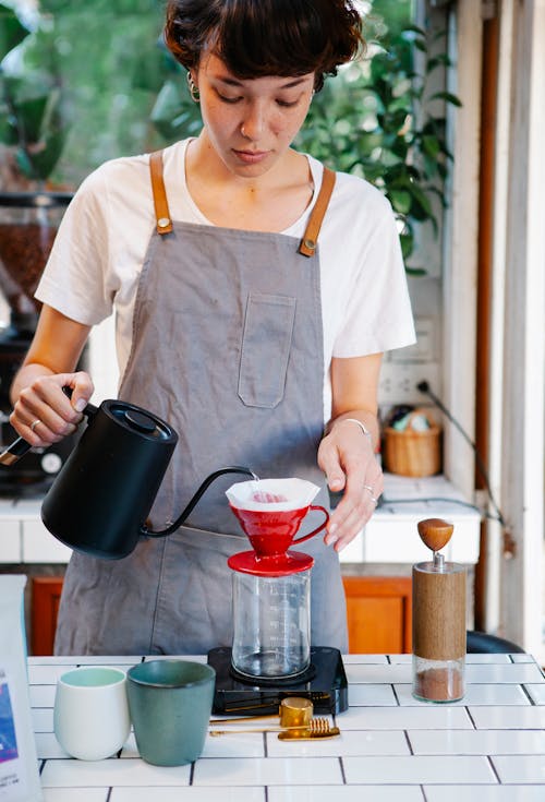 Crop female barista in gray apron standing at table and preparing coffee in alternative way in cafe in daytime