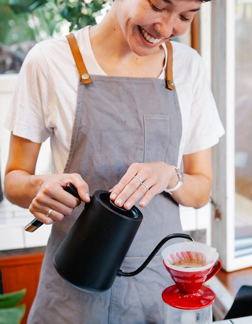 Crop positive female in apron preparing coffee using pour over coffee maker and drip kettle in light cafe