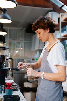 Focused woman barista in gray apron standing and grinding coffee beans with manual stainless steel grinder for pour over coffee in cafe