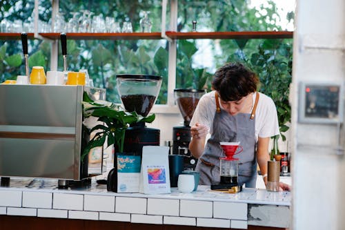 Thoughtful female barista with brown hair in apron pouring aromatic alternative coffee while working in light cafe in daytime