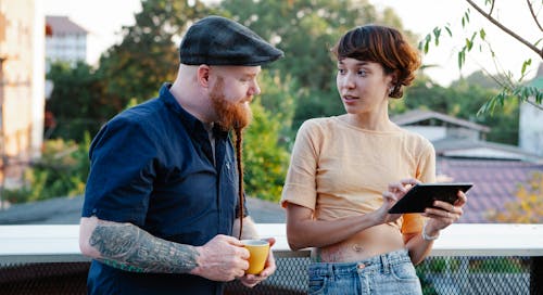 Young man with beard in cap and woman using tablet speaking on terrace in daytime in summer