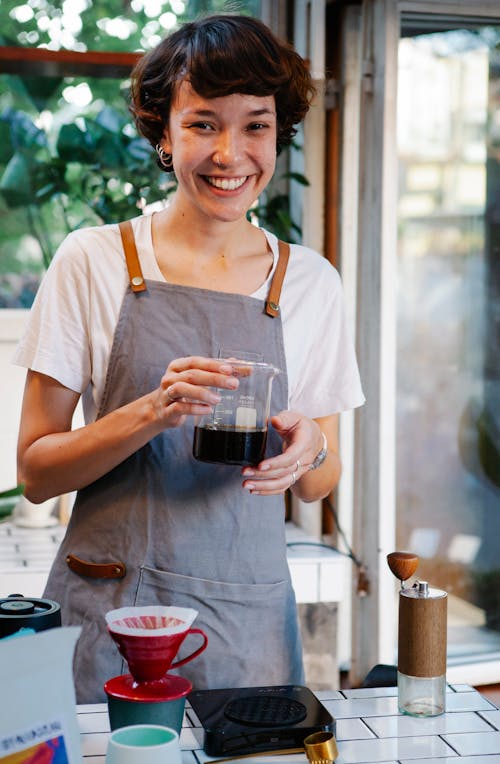 Young positive female smiling and looking at camera while preparing tasty aromatic coffee in kitchen of cafeteria in daytime