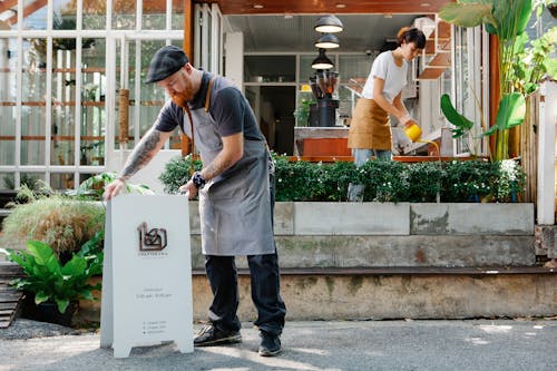 Man in cap and apron installing signboard on street while woman watering verdant plants in cafe