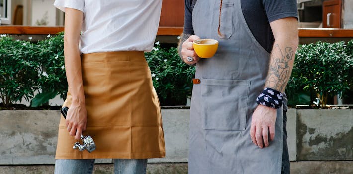 Two baristas in aprons holding coffee and espresso tool outdoors in a casual urban setting.