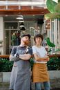 Serious man and woman wearing aprons standing on terrace of modern coffee shop with filter holders in hands on street