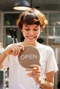 Through glass of happy female worker holding wooden Open sign working in modern cafe with glowing lamp inside