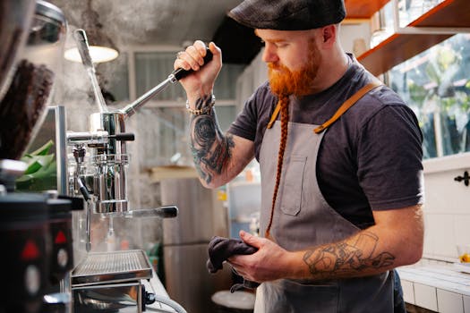 Side view of tattooed male with beard wearing cap standing near coffeemaker with lever and steam while working in coffee house