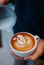 Unrecognizable man pouring hot milk from pitcher into cup of latte with creative pattern while working in coffee house on blurred background