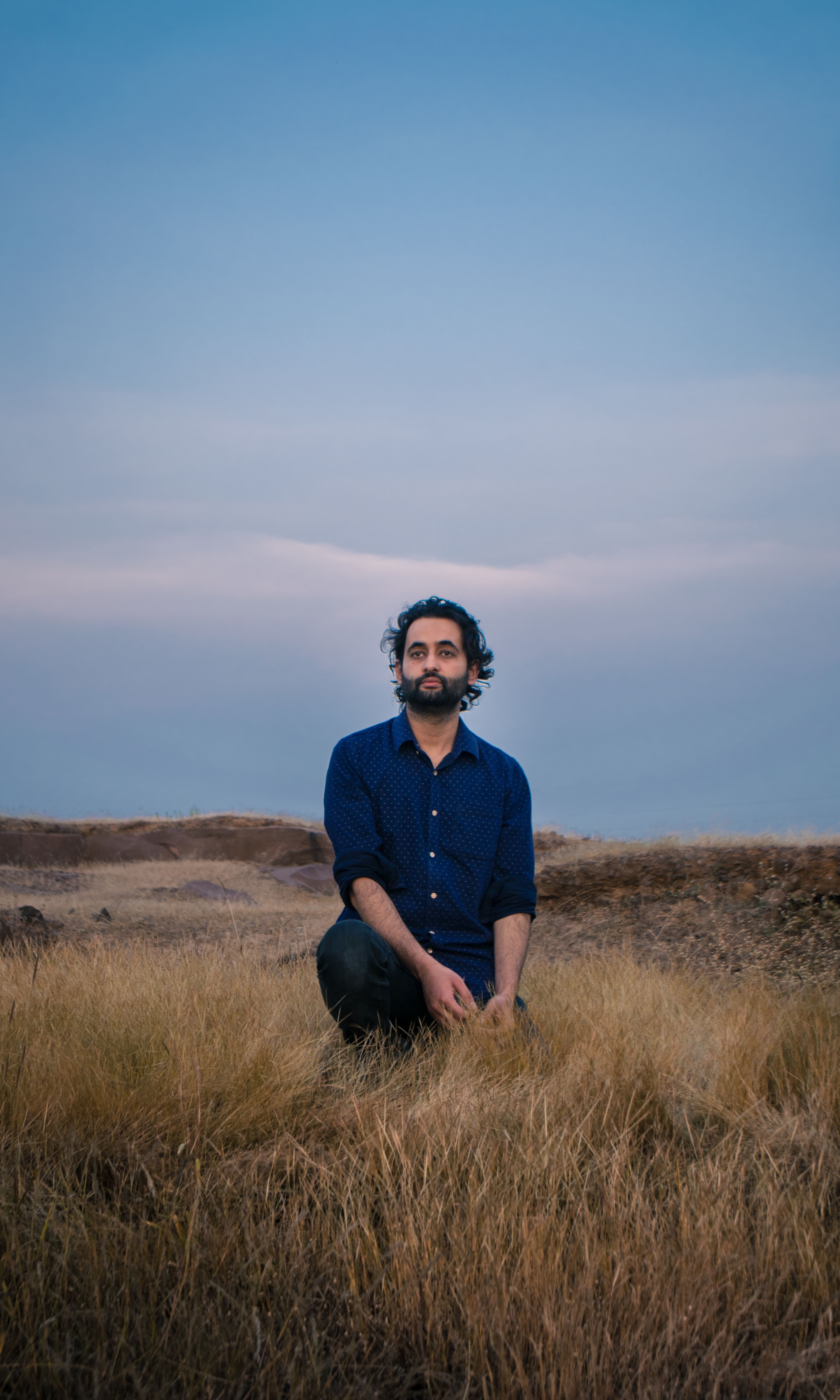 Bearded man in blue shirt seated in a grassy field during twilight, calm and contemplative.