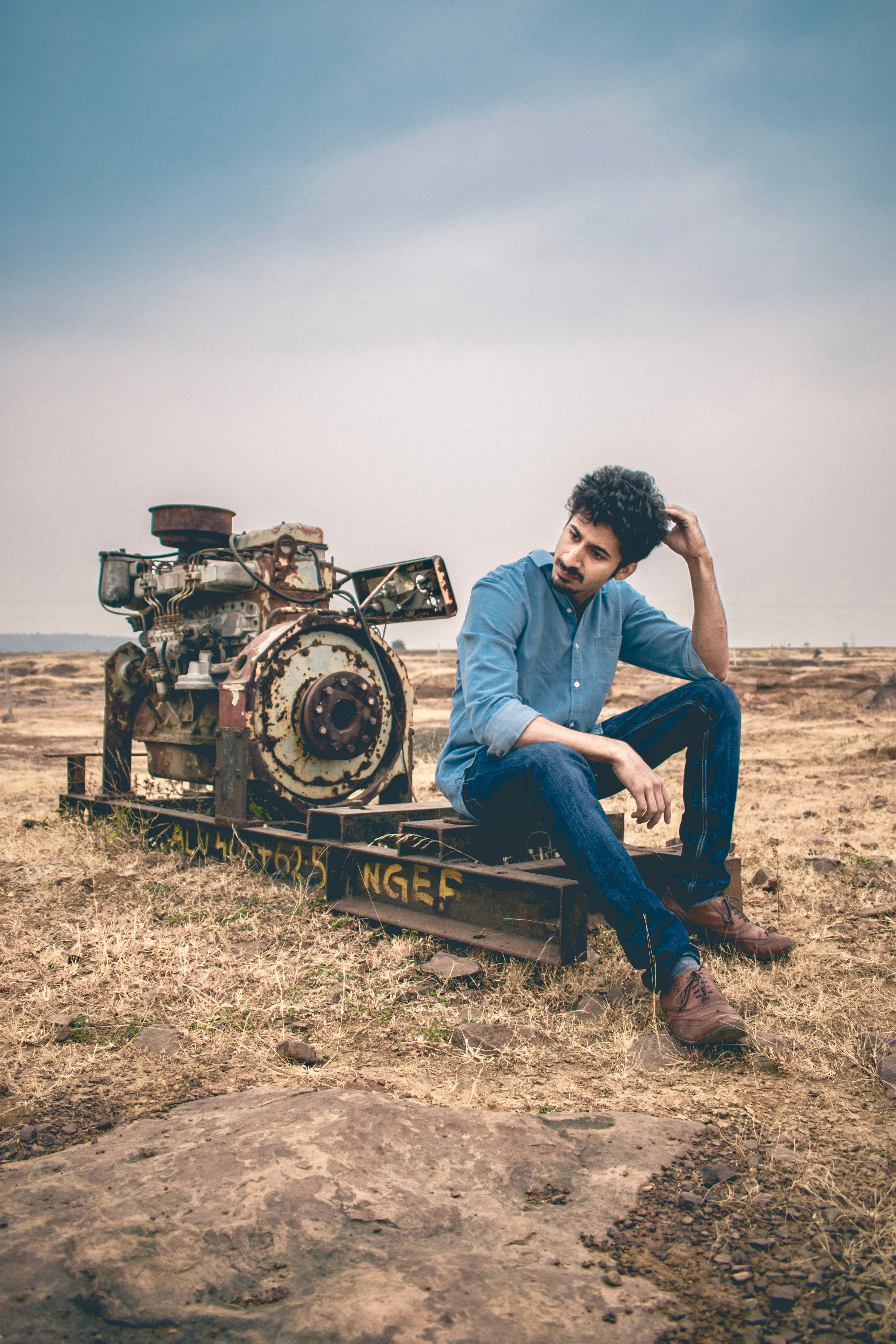 A man in a blue shirt sits thoughtfully beside rusted machinery in an open field.
