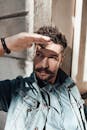 Pondering male with mustache wearing denim jacket covering eyes by hand from bright sunlight and looking away in deep thoughts