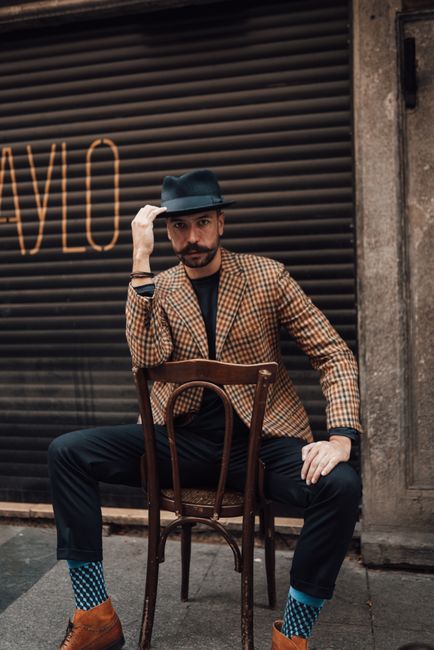 Confident handsome male in classy trendy suit touching hat brim and looking at camera while sitting on chair backwards against garage wall