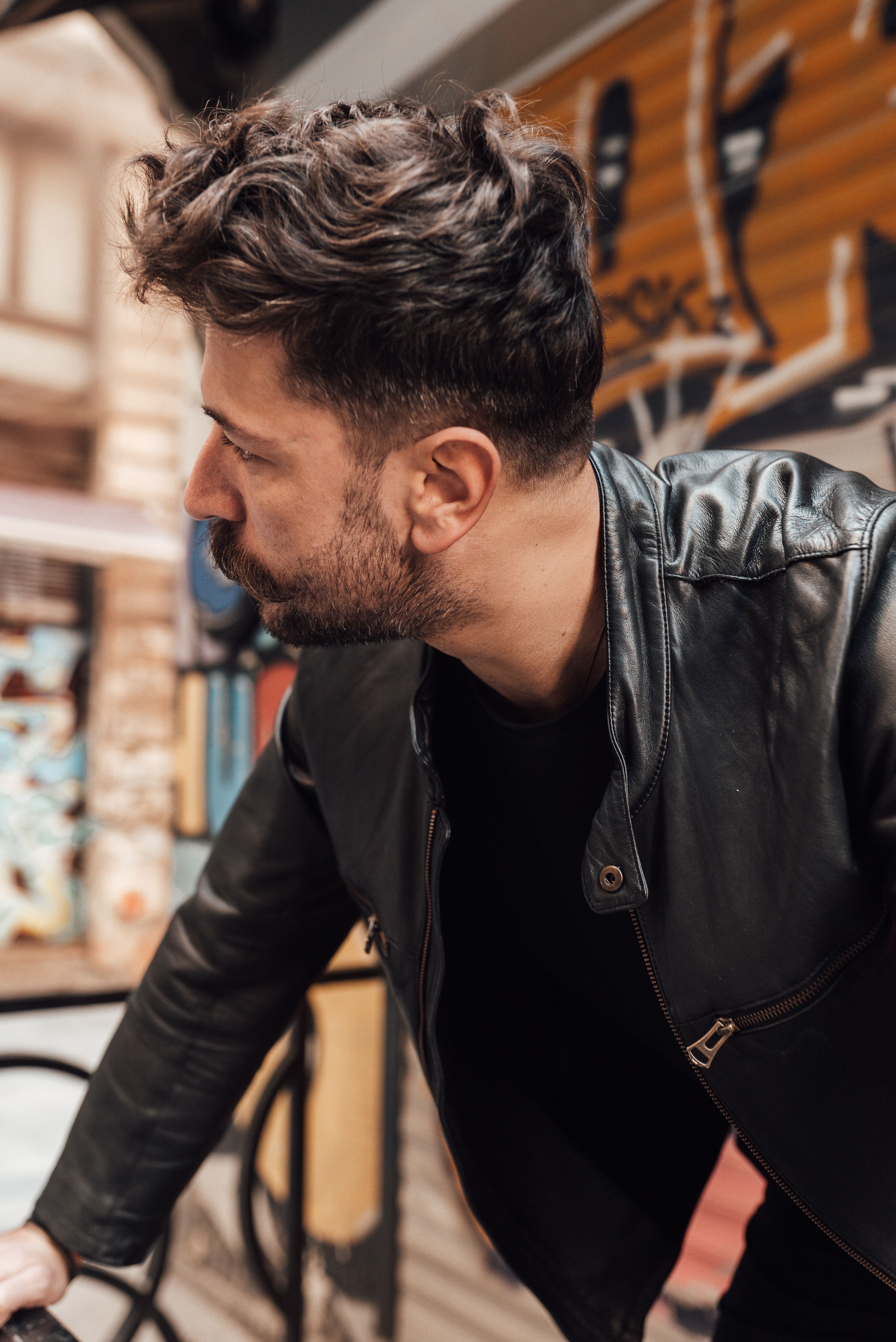 Contemplative bearded male in black leather jacket standing on street with vandalized building walls and looking away pensively