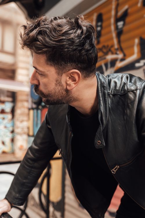 Free Contemplative bearded male in black leather jacket standing on street with vandalized building walls and looking away pensively Stock Photo