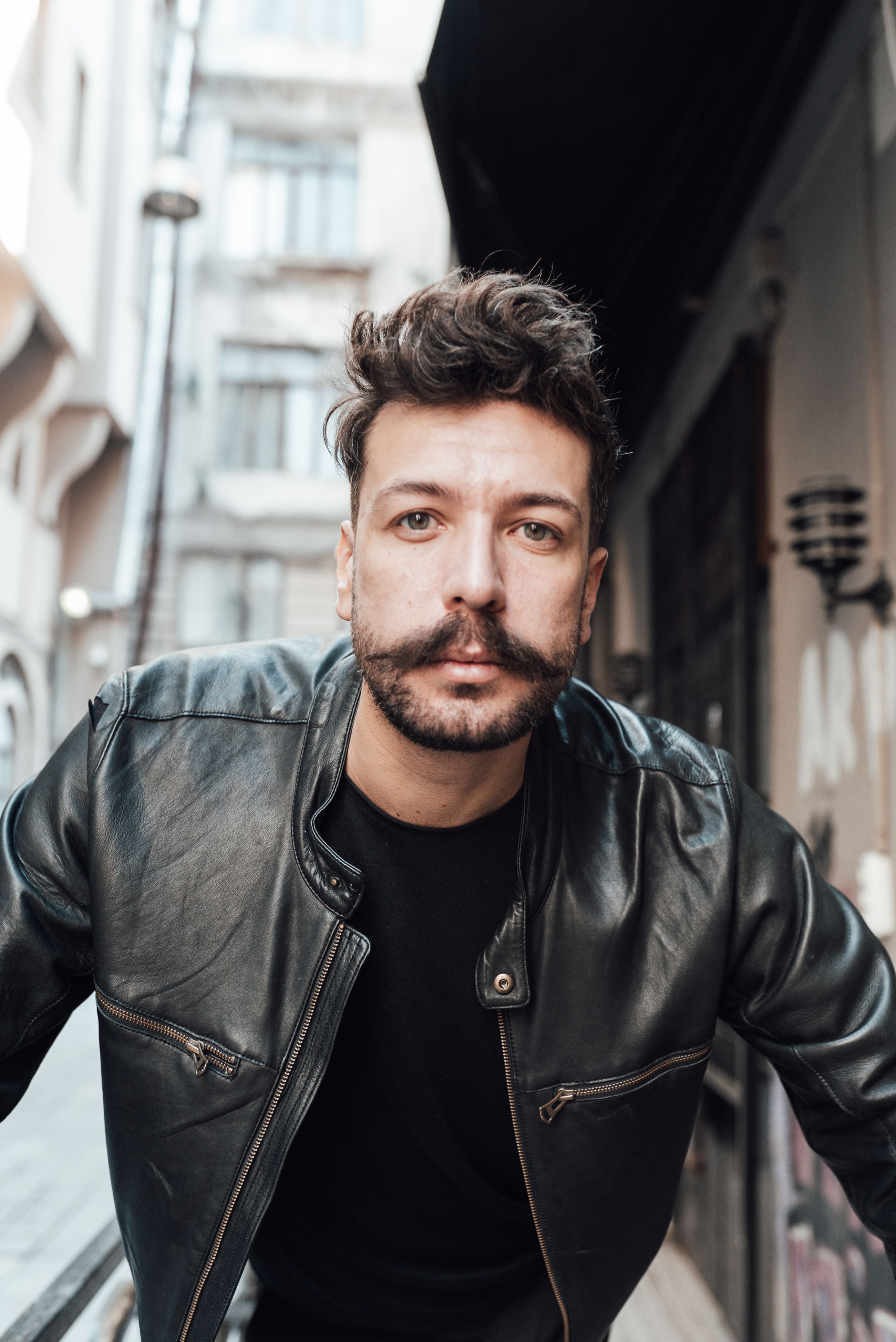 Calm adult male with mustache and beard wearing black leather jacket standing on modern city street and looking at camera