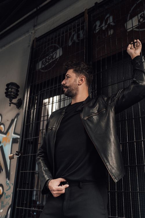 Free Serious adult bearded male in black leather jacket with arm raised and hand on pants fly standing near grid wall and looking away Stock Photo