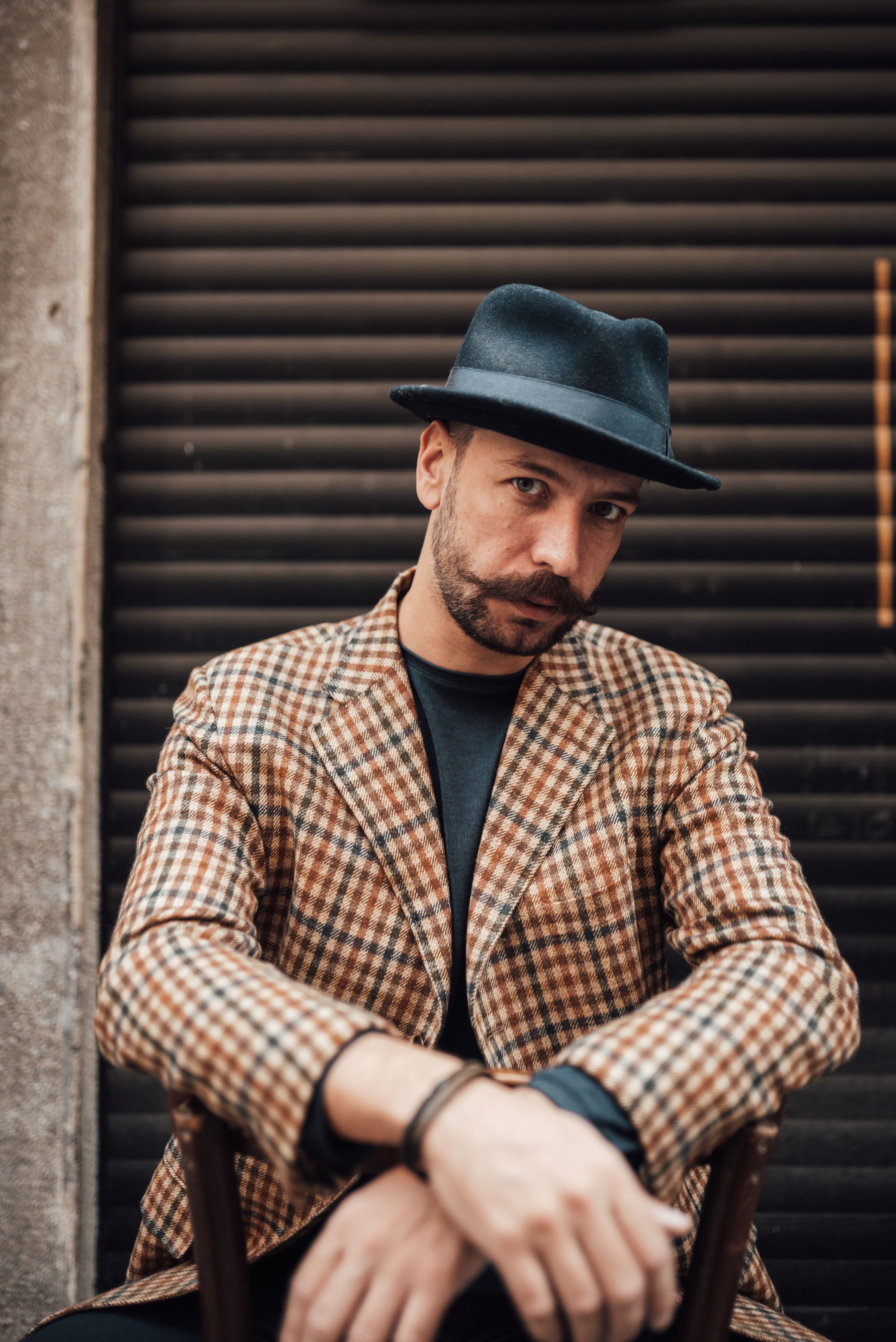 Confident well dressed adult male with mustache wearing trendy checkered jacket and hat sitting on chair backwards against garage wall and looking at camera