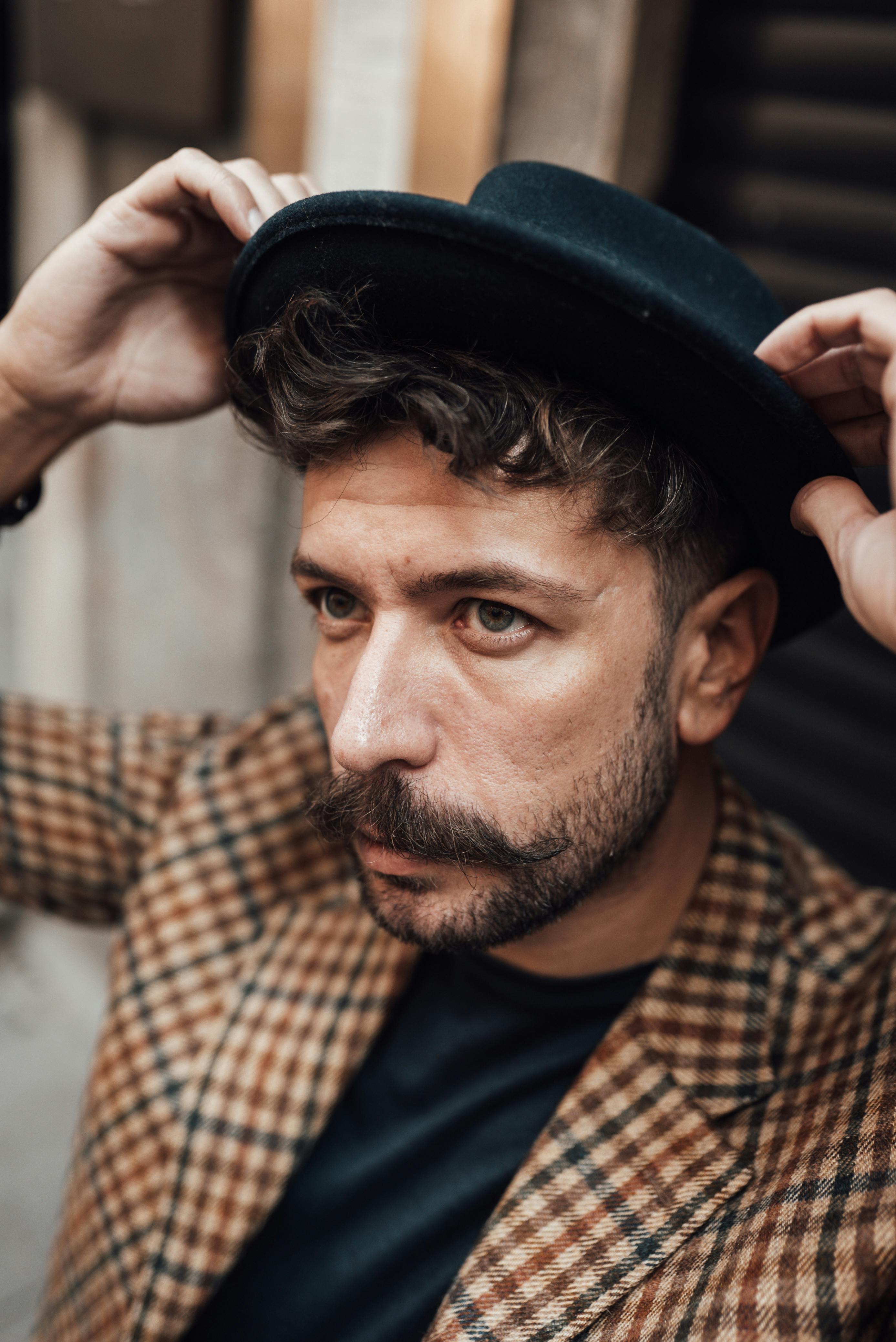 Thoughtful handsome male with mustache in trendy checkered jacket putting on black hat and looking away on street