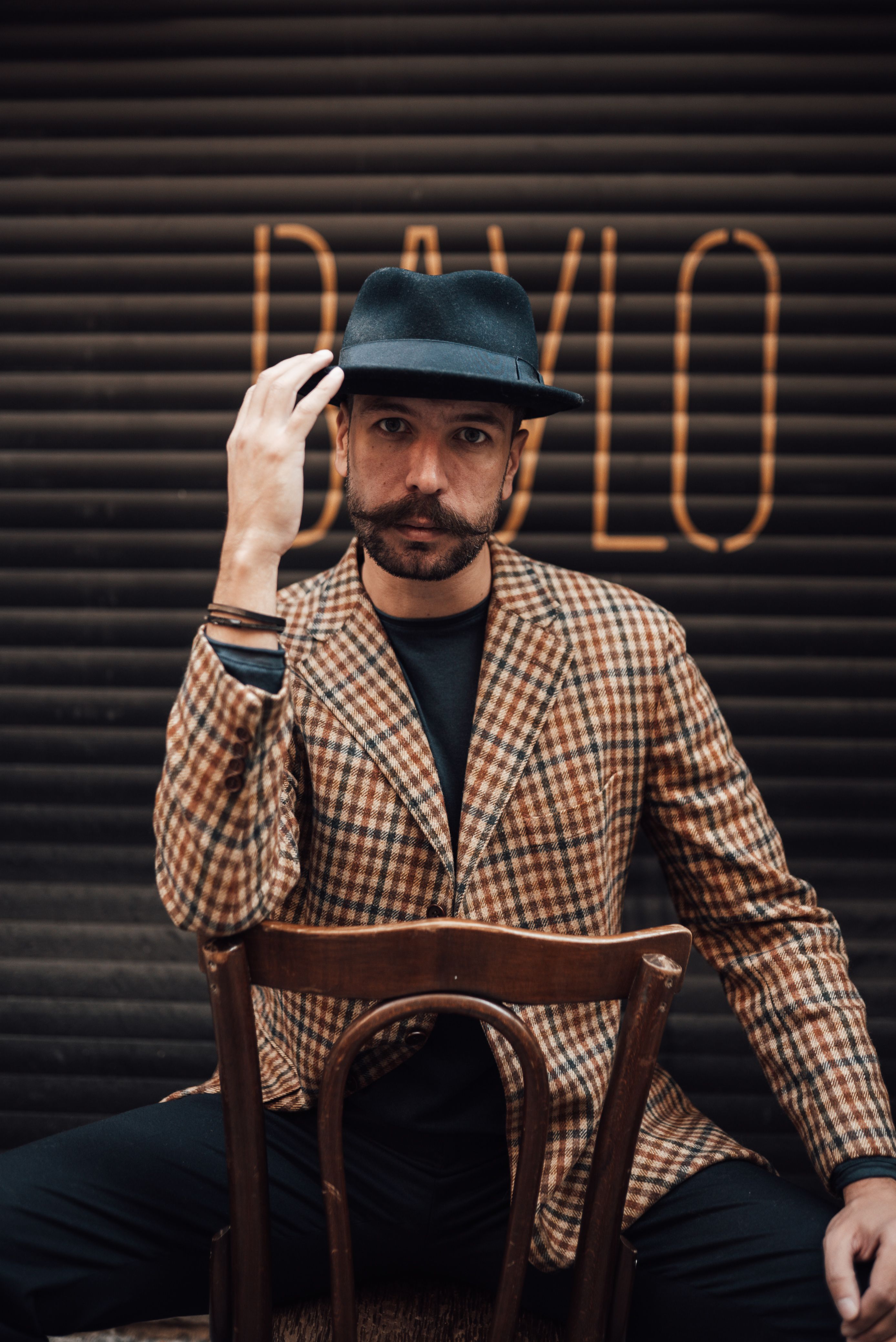 Stylish bearded male with mustache wearing posh suit touching hat and looking at camera while sitting on wooden chair backwards against garage wall