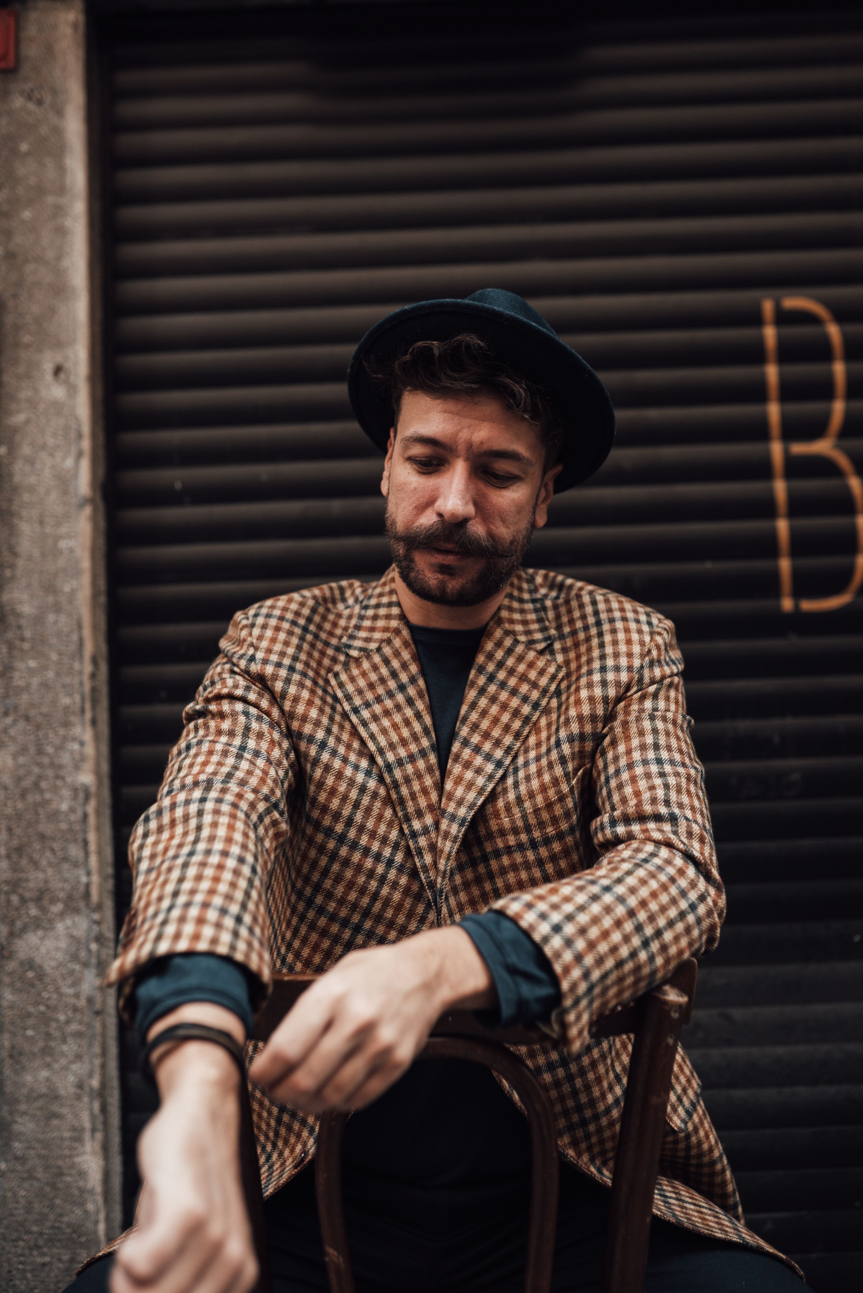 Content adult bearded male in posh suit and hat sitting on wooden bench and putting on bracelet against garage wall