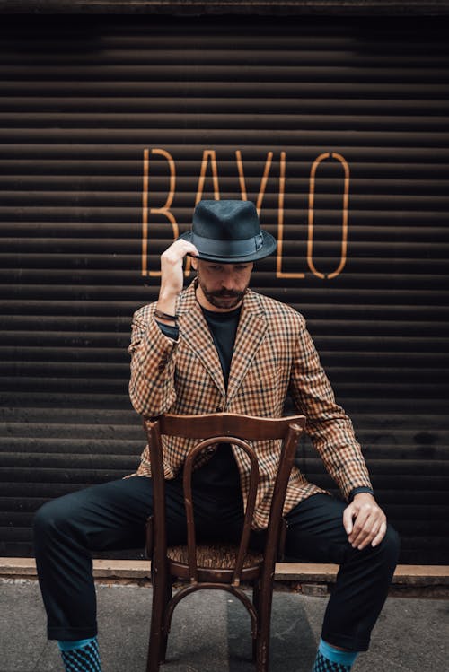 Stylish pensive adult male with mustache and beard touching trendy hat and looking down while sitting on chair backwards against garage wall