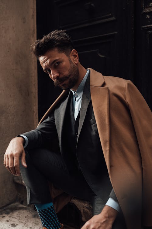 Confident well dressed adult male in classy black suit and coat sitting on old house doorway and looking at camera calmly