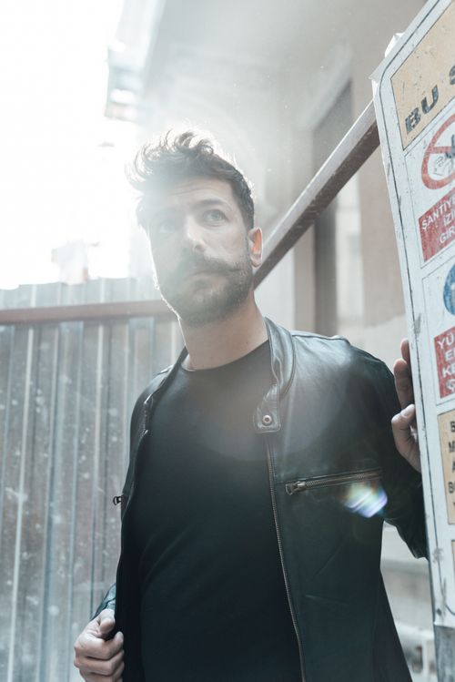 Free From below of pensive man with beard and mustache in black outfit standing on street and looking away in daytime Stock Photo