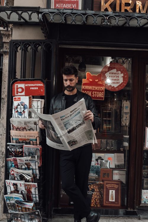 Free Thoughtful man with dark hair and beard in trendy black outfit standing on street and reading newspaper against counter of book store in daytime Stock Photo