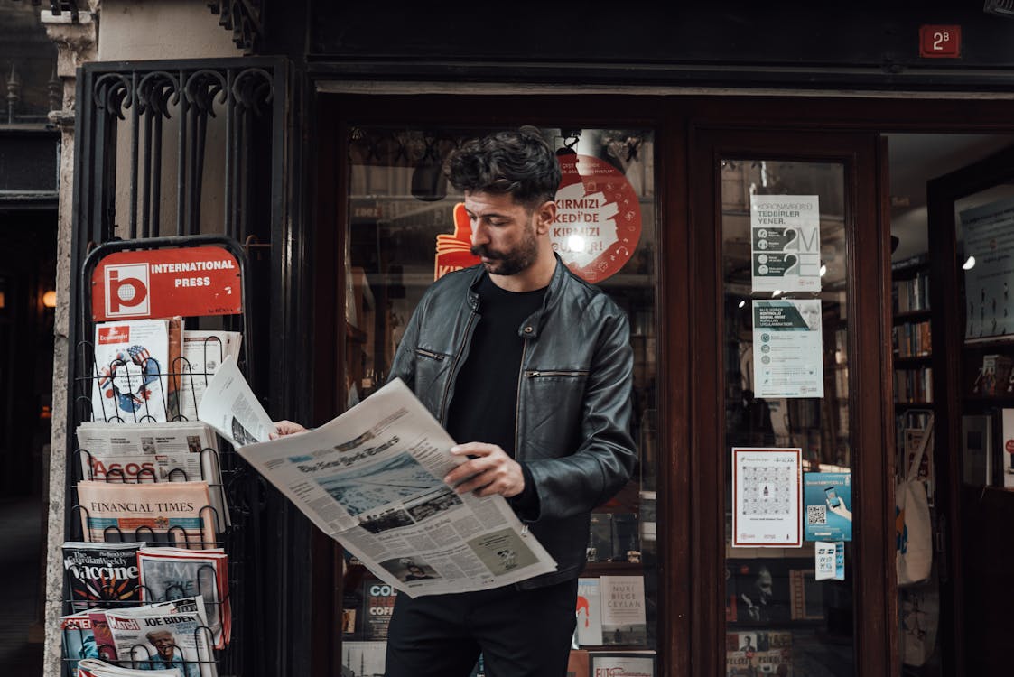 Serious bearded male with trendy haircut in casual clothes reading newspaper while standing near entrance of shop