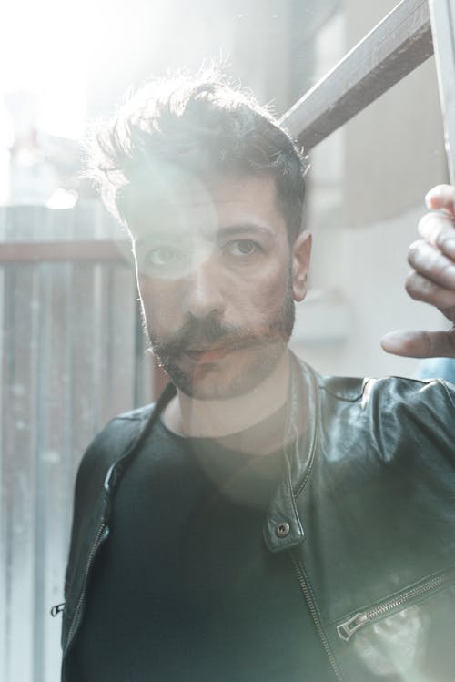 Confident male with mustache and trendy hairstyle standing near metal fence under bright sunlight and looking away