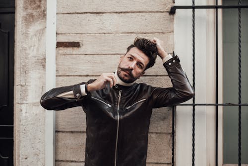 Stylish male hipster with trendy haircut and mustache in black leather jacket touching hair while looking at camera