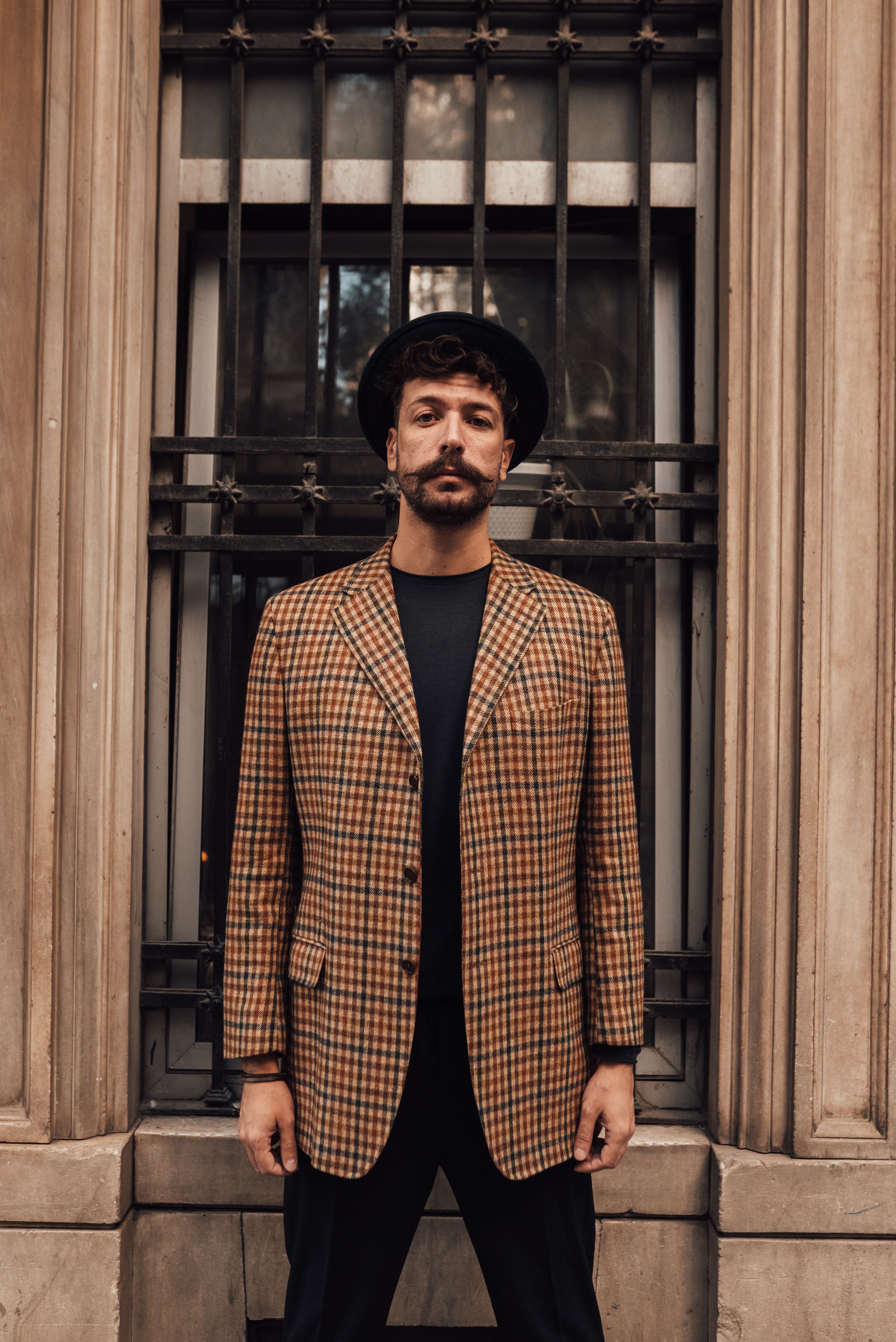 Confident male model with beard in trendy outfit looking at camera while standing near building